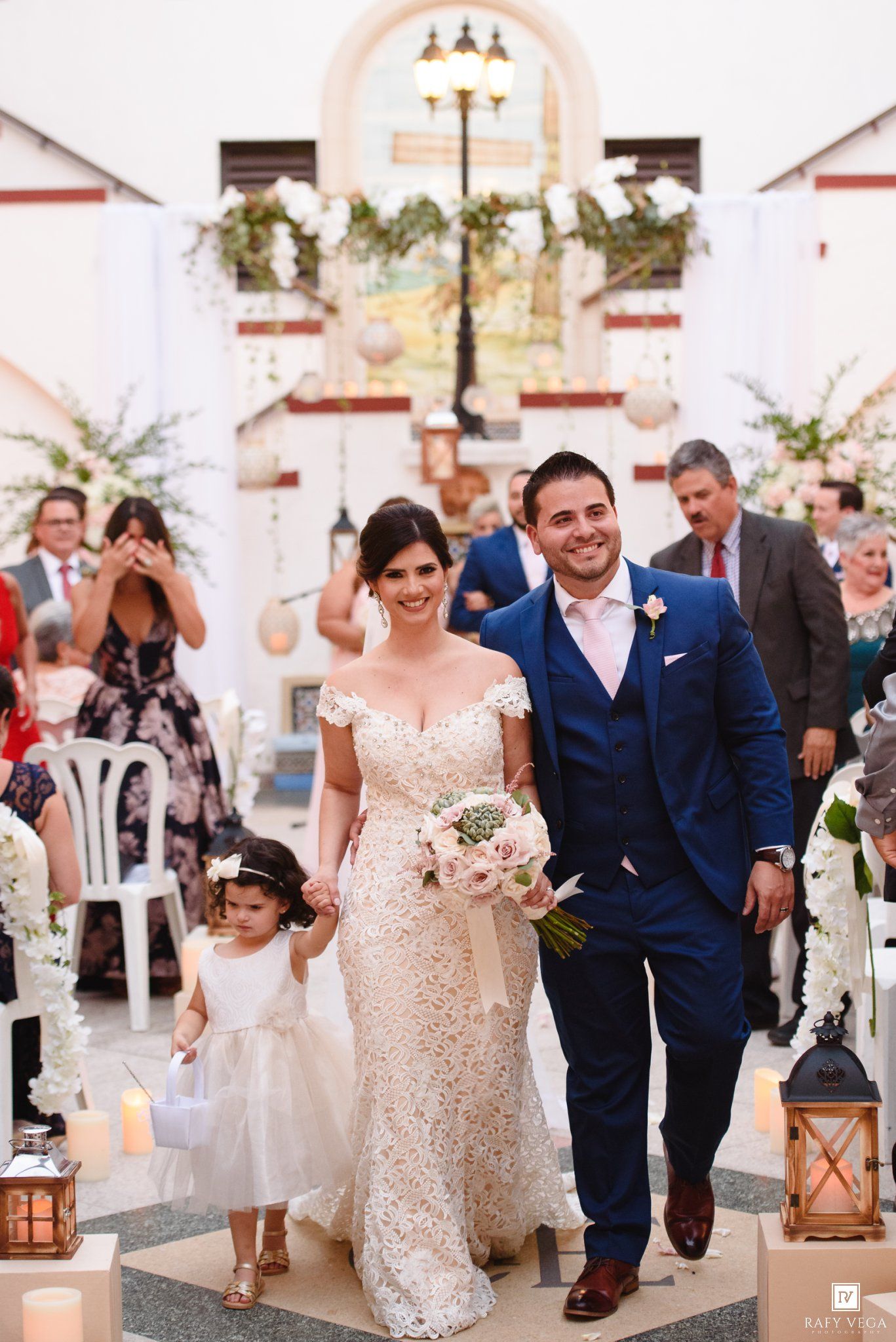 Bride in lace gown and groom in blue suit walk down church aisle with flower girl after wedding ceremony.