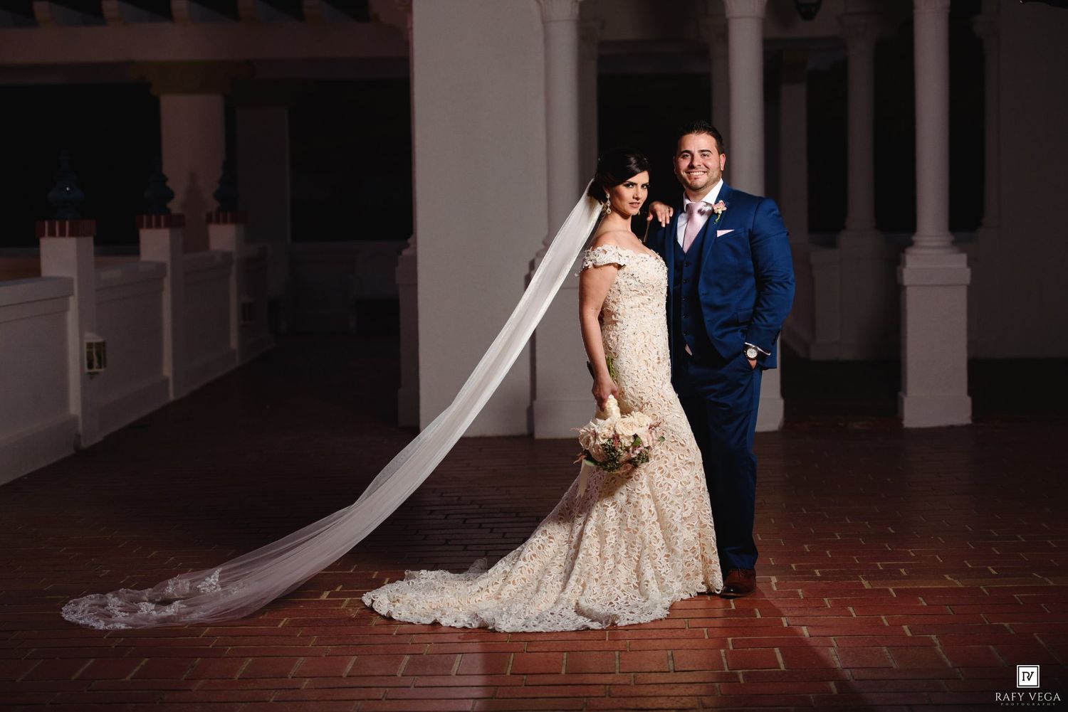 Bride in lace mermaid gown with long veil stands beside groom in navy suit on brick venue floor.