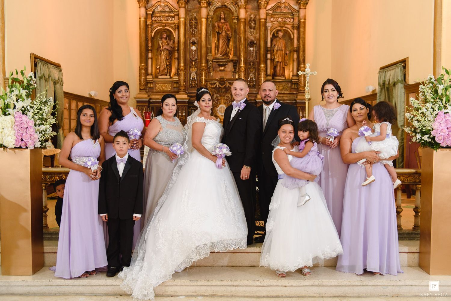 Wedding party poses in church with bride, groom, bridesmaids in lavender gowns, and children near ornate golden altar.