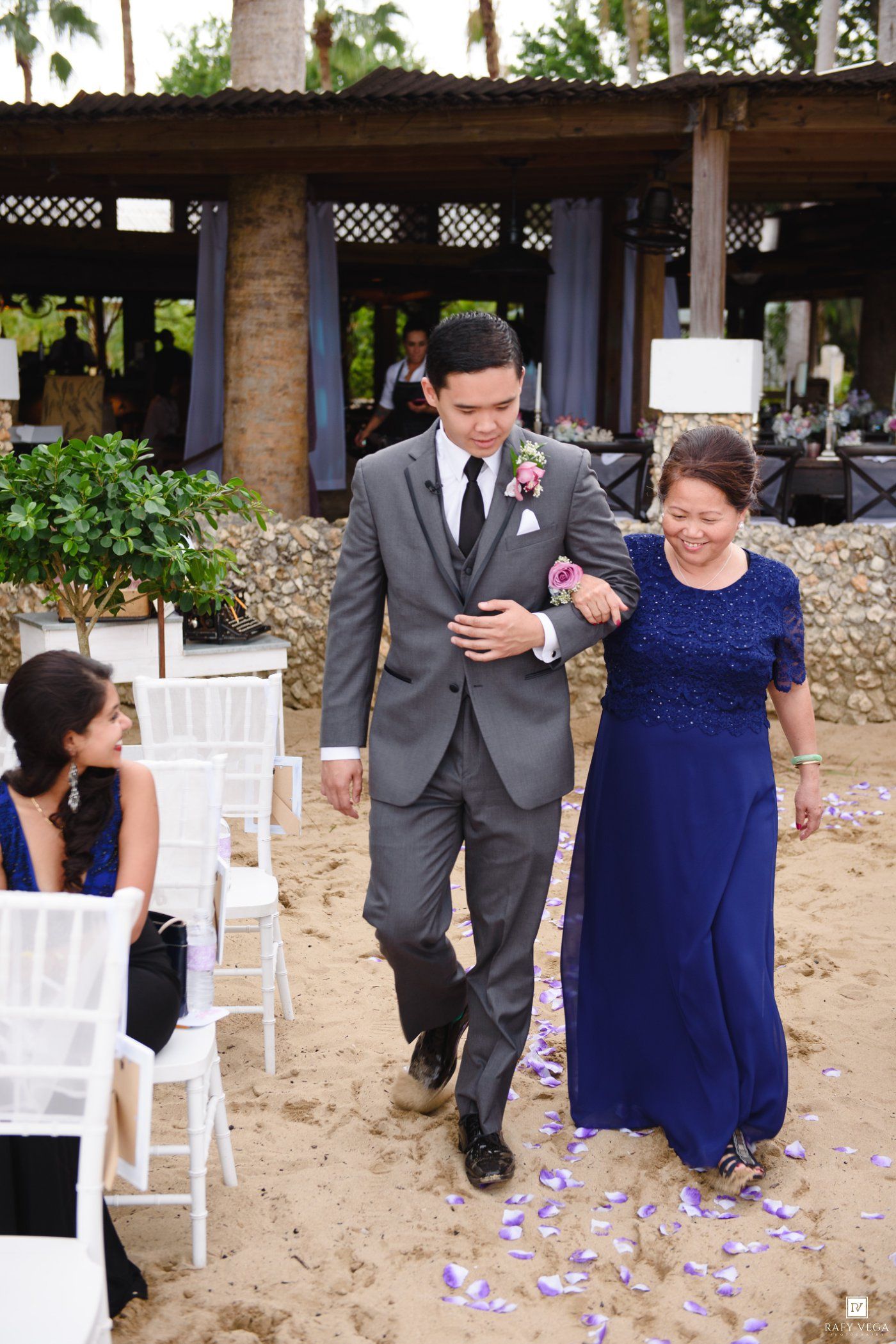 Groom in gray suit walks down sandy aisle with mother in blue gown at outdoor beach wedding ceremony.