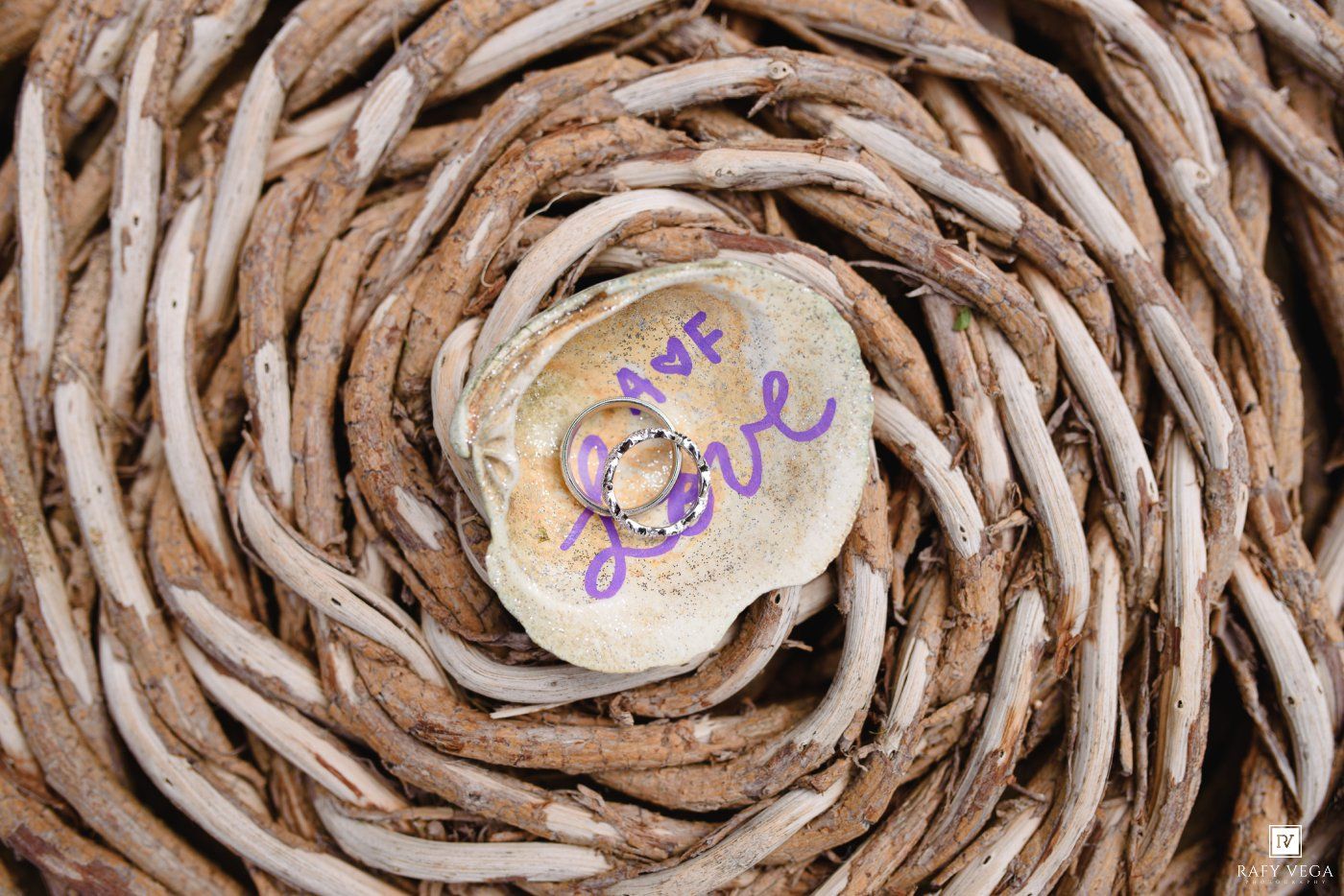 Two gold wedding rings nestled in a shell inscribed with love, placed in a woven wicker spiral centerpiece.