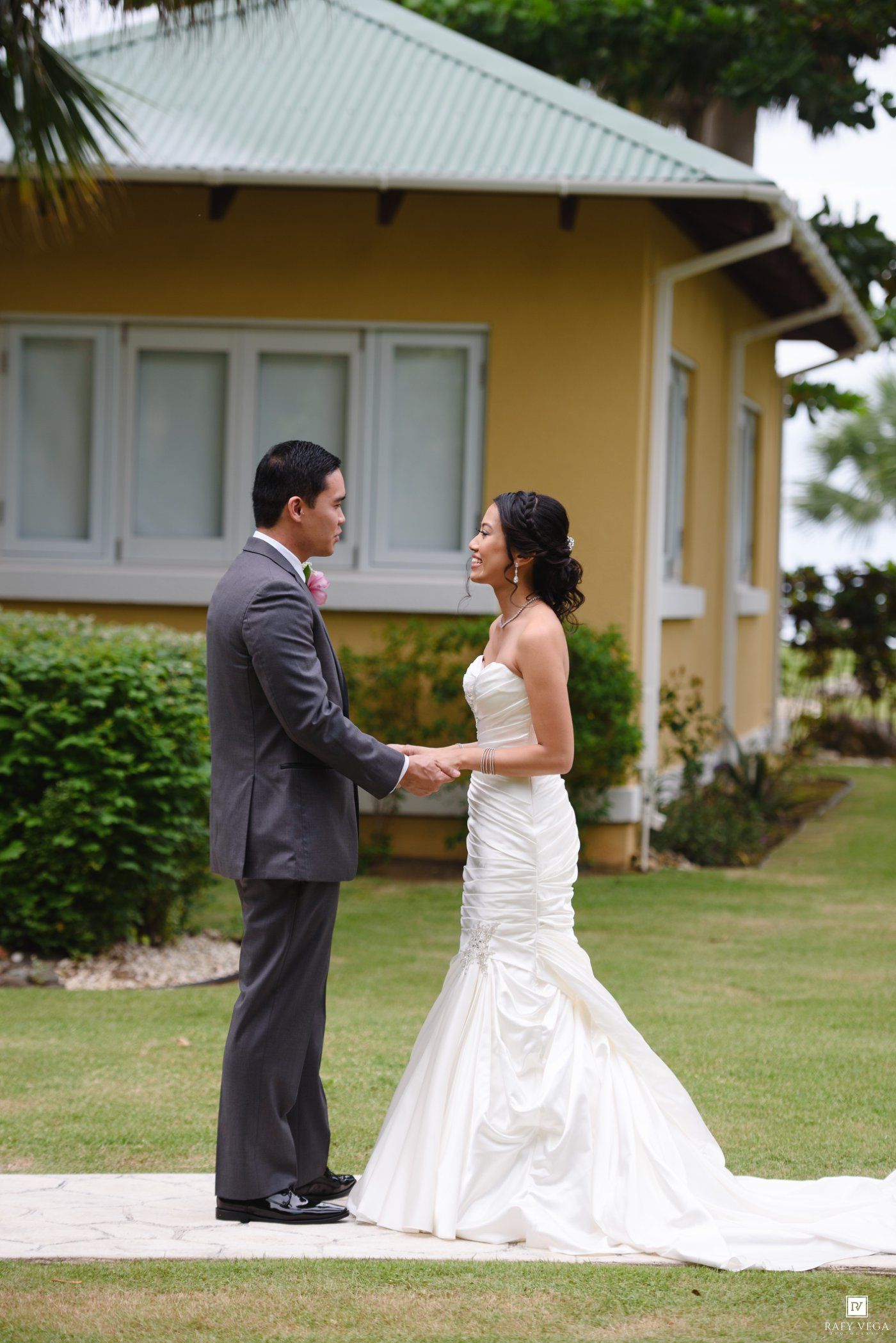 A bride and groom hold hands and share a tender moment on a green lawn outside a yellow venue.