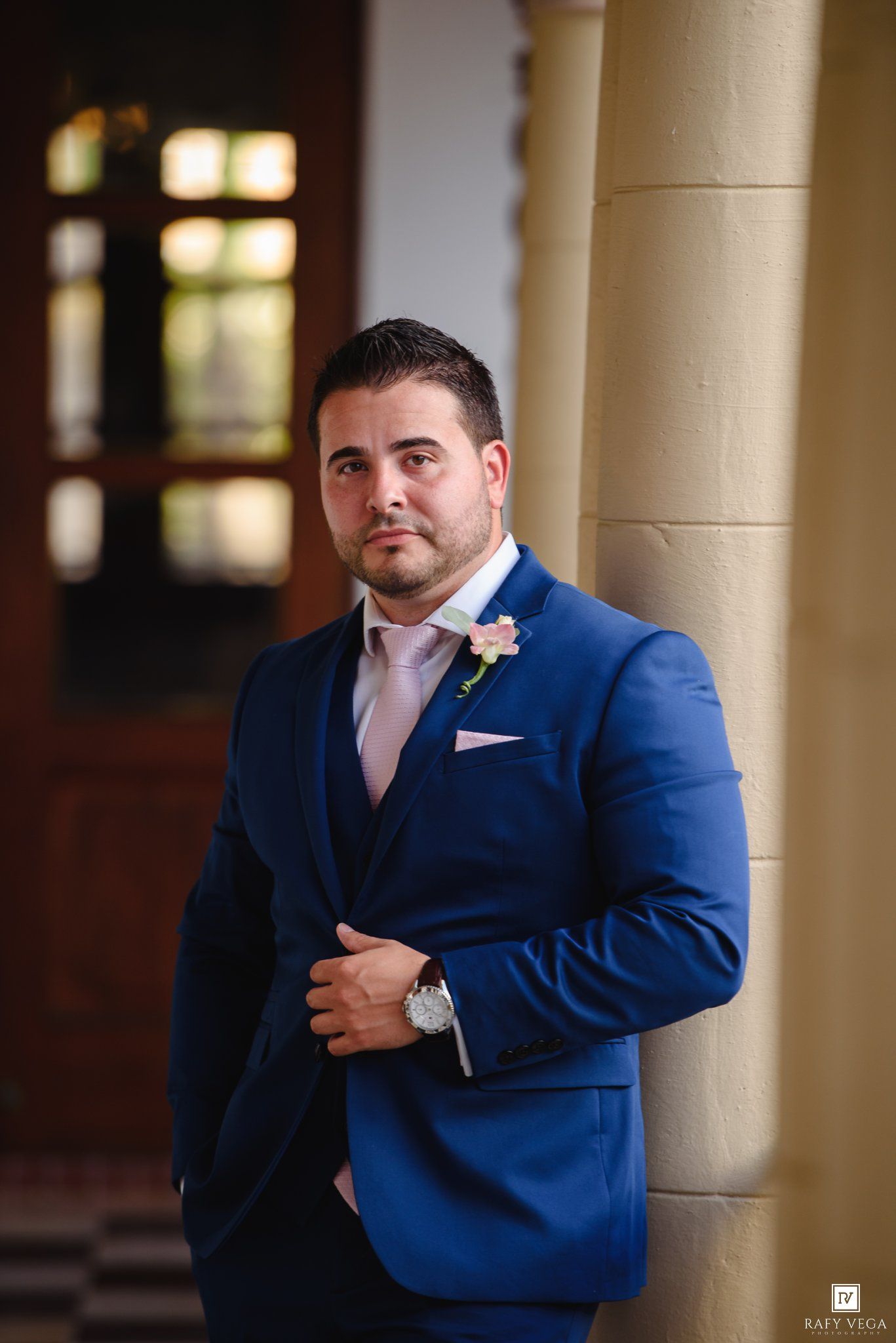 Handsome groom in navy blue three-piece suit with pink tie and boutonniere poses at historic wedding venue.