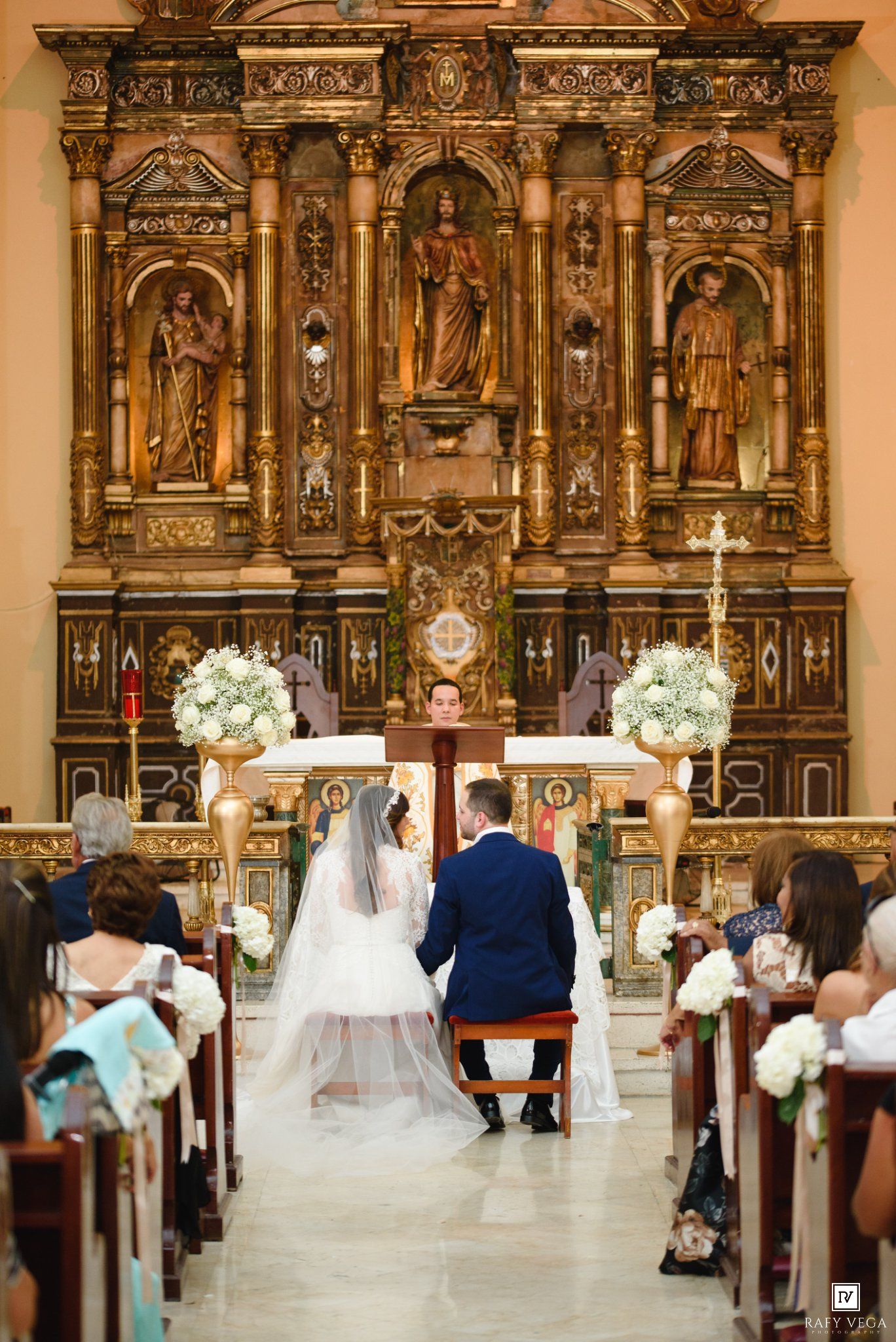 Bride and groom kneeling at ornate golden Catholic church altar during wedding ceremony with white floral arrangements.
