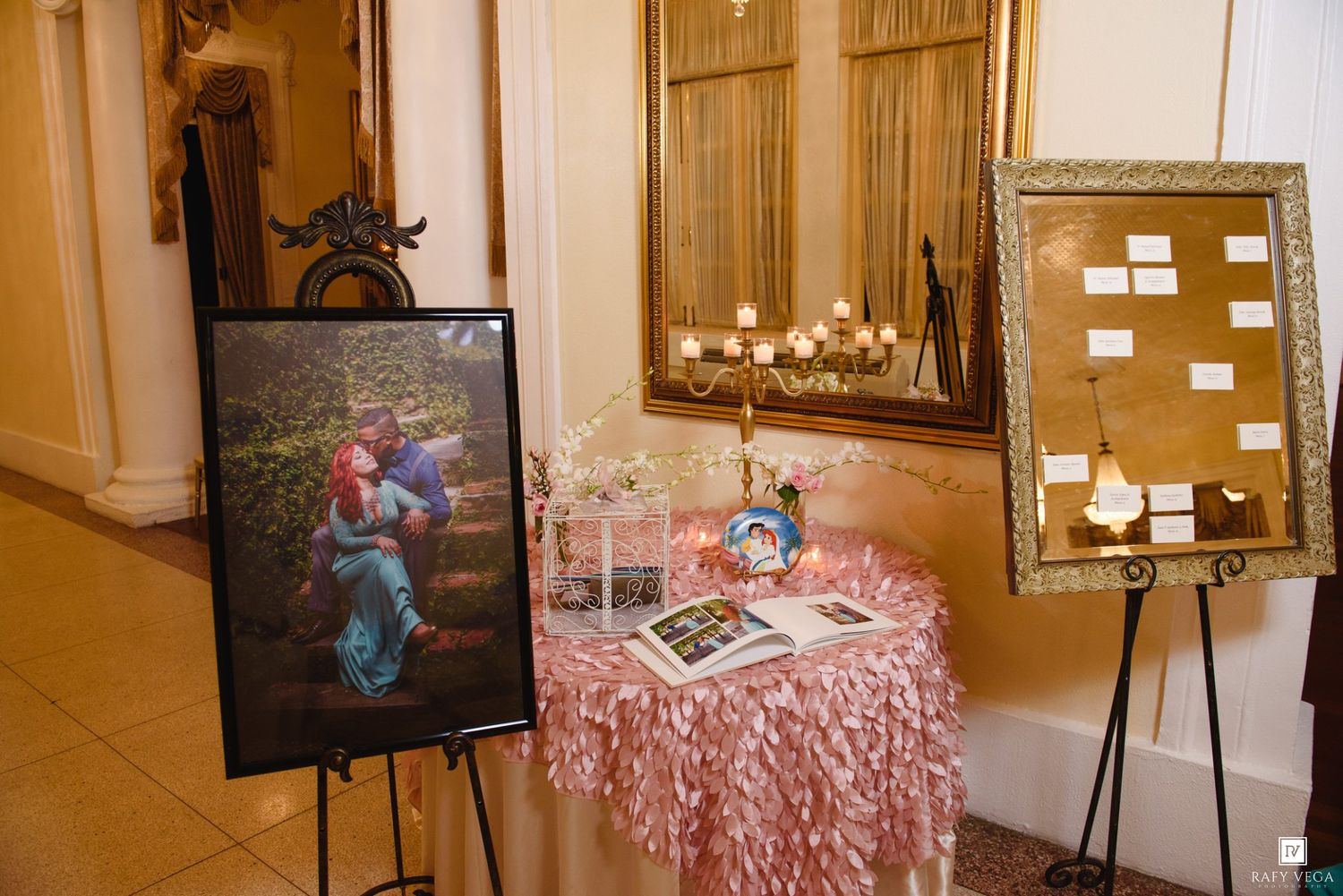 Elegant wedding reception display with framed photo, pink ruffled table, seating chart on gold easel, and candles in ornate venue.