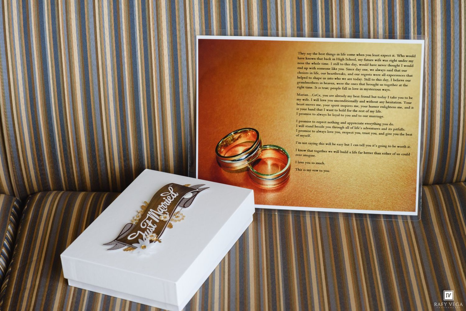 Two gold wedding rings displayed on a printed card beside a white gift box on a striped fabric surface.