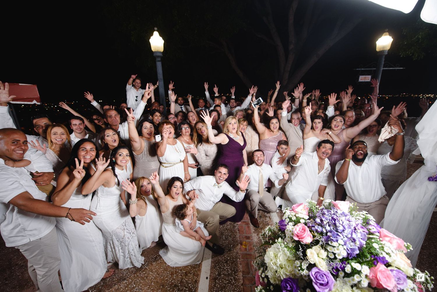 Large group of people dressed in white celebrating outdoors at night, raising hands joyfully near a lamp post.