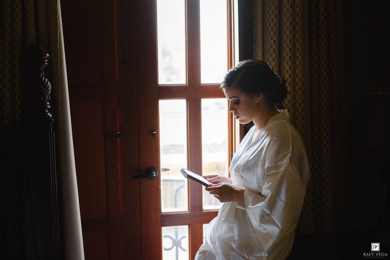 Bride in white robe reading a card by a sunlit window on her wedding morning.