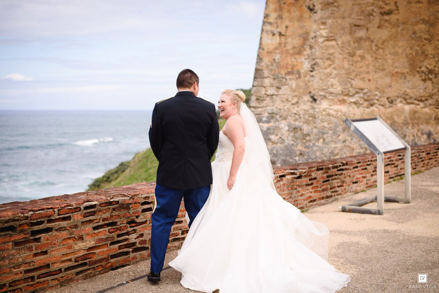 Bride and groom stand together overlooking the ocean at a historic brick fort, bride in white gown with veil.