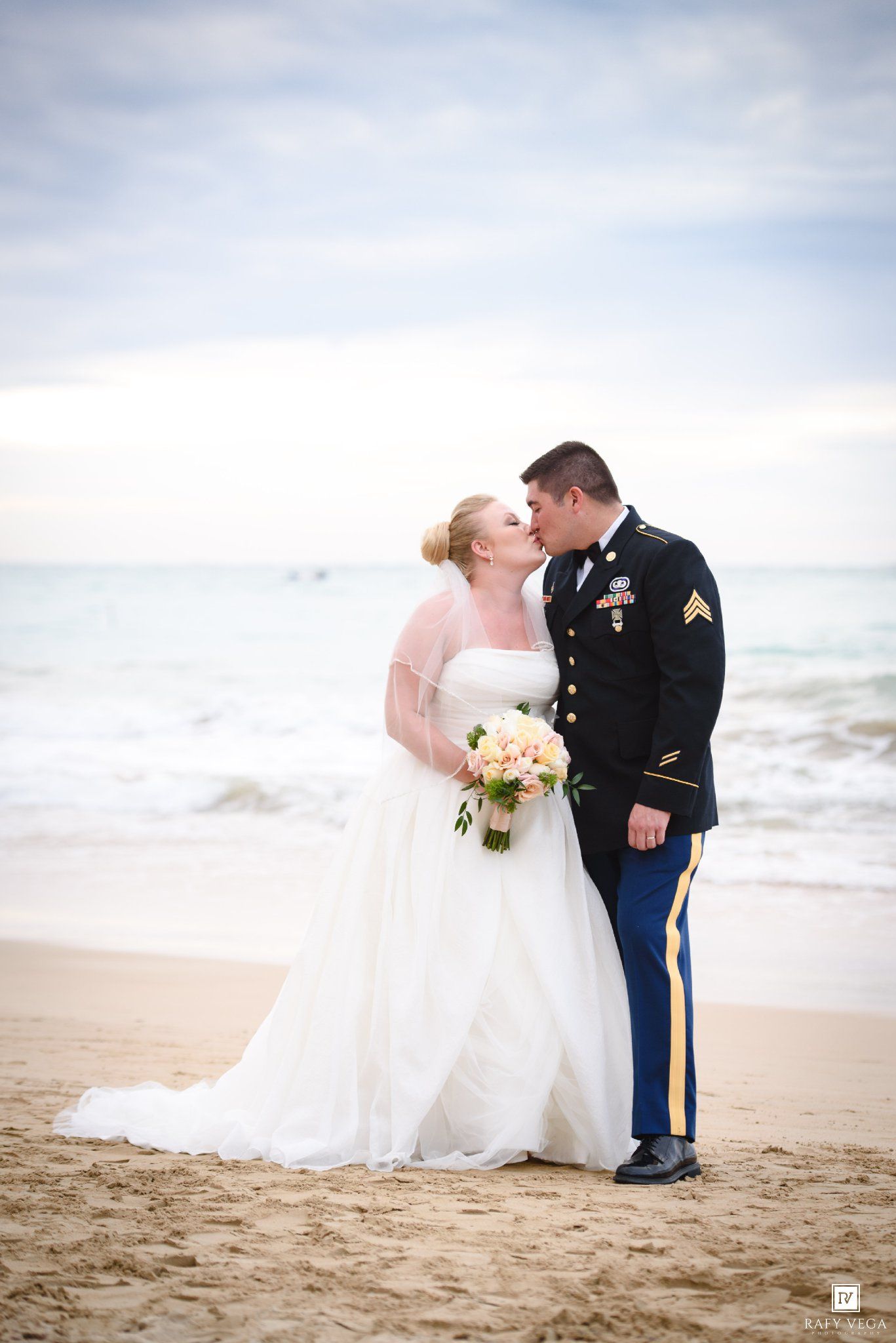 Military groom in dress uniform kisses bride in white gown holding floral bouquet on sandy beach with ocean backdrop.
