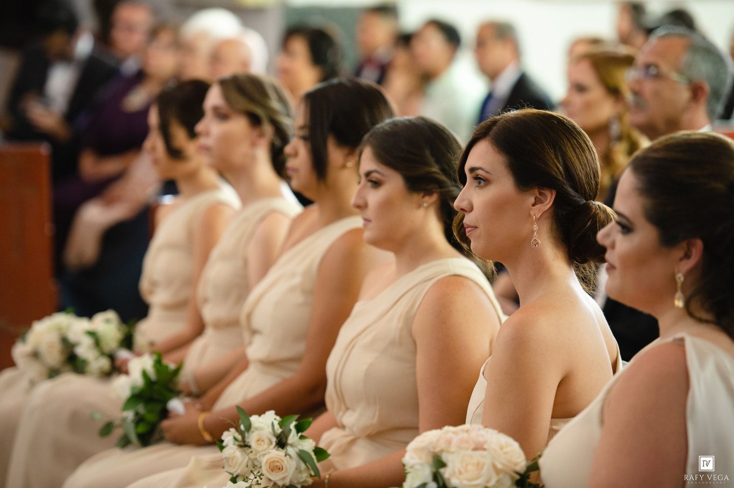 Bridesmaids in matching champagne gowns holding white bouquets seated at a wedding ceremony.