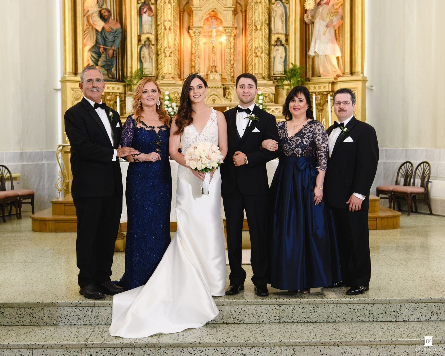 Wedding party poses with bride and groom before golden church altar in formal attire.