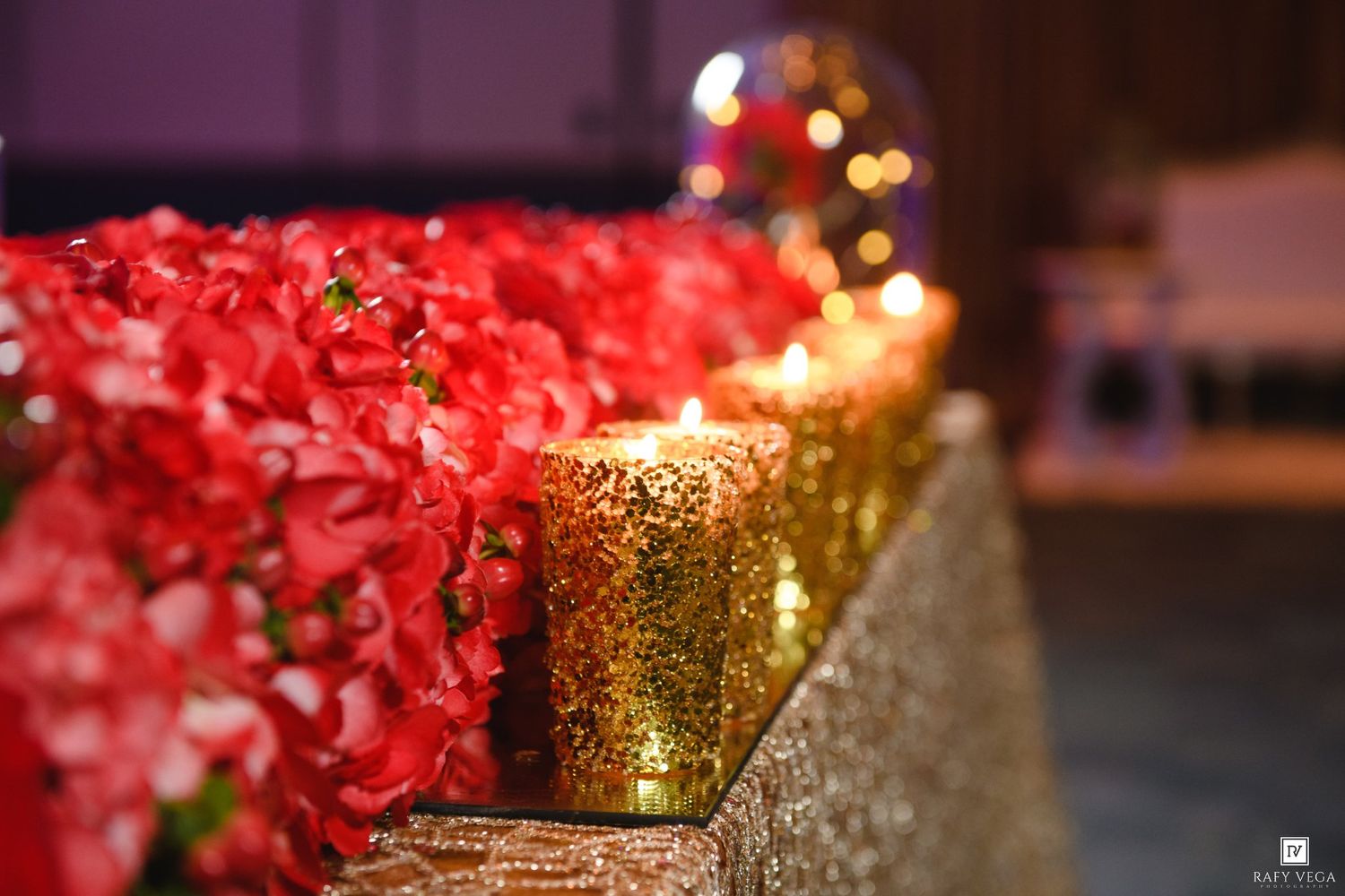 Red hydrangeas and gold glittery candle holders line a wedding aisle, with bokeh lights in the background.