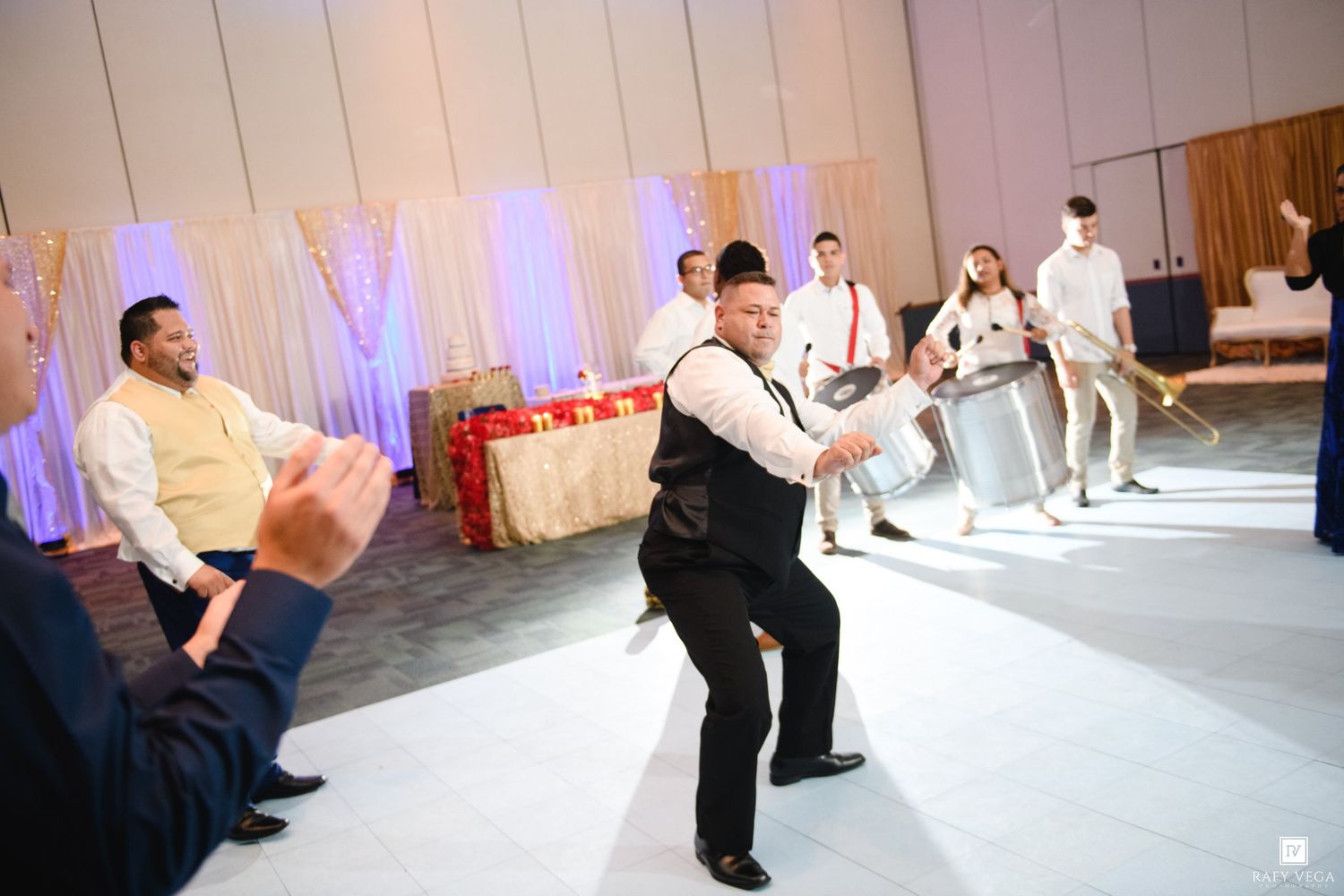 Energetic wedding reception dance performance with guests clapping on a white dance floor in an elegant ballroom.