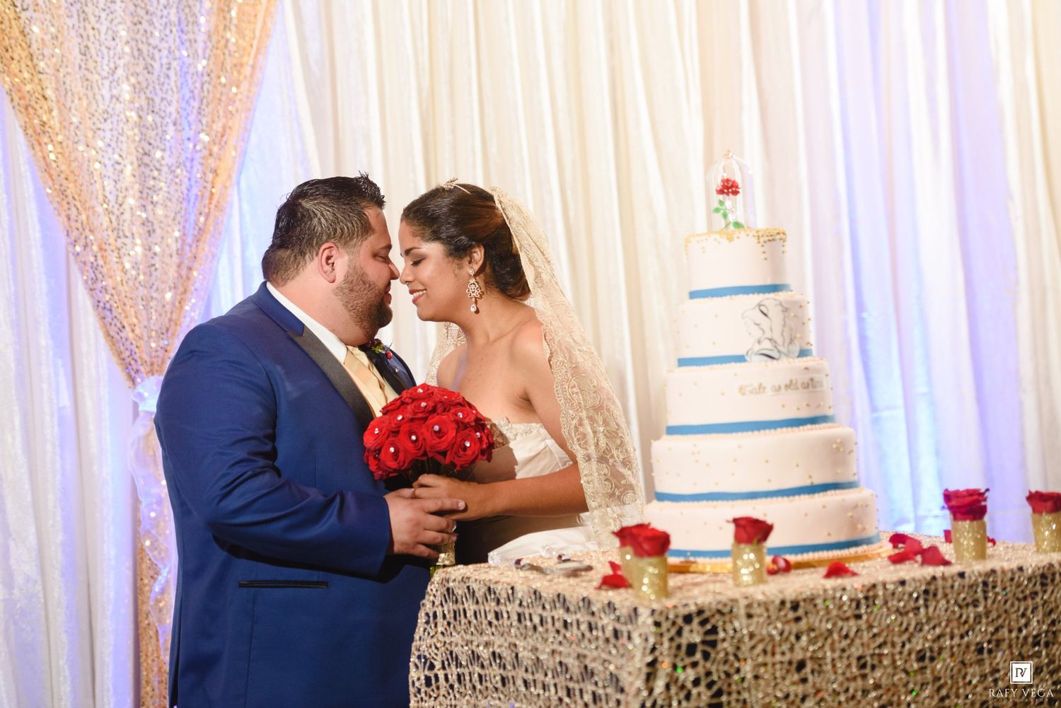 Bride and groom sharing a romantic moment beside a white wedding cake adorned with red roses at their reception.
