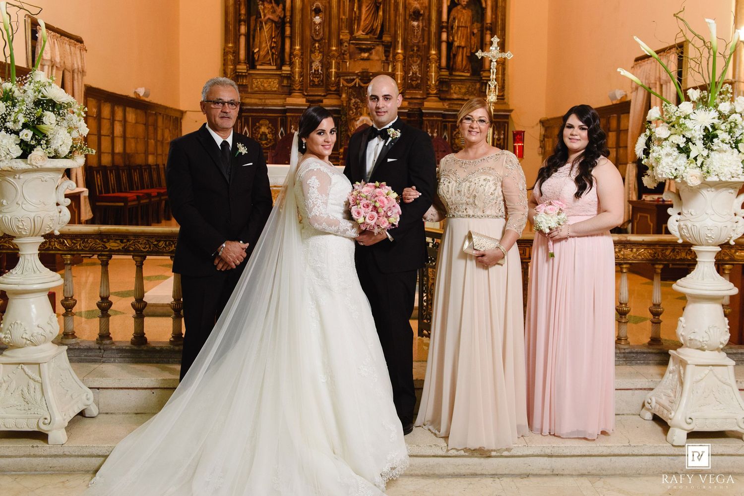 Wedding party poses at church altar with bride, groom, and family in elegant pink and white attire.