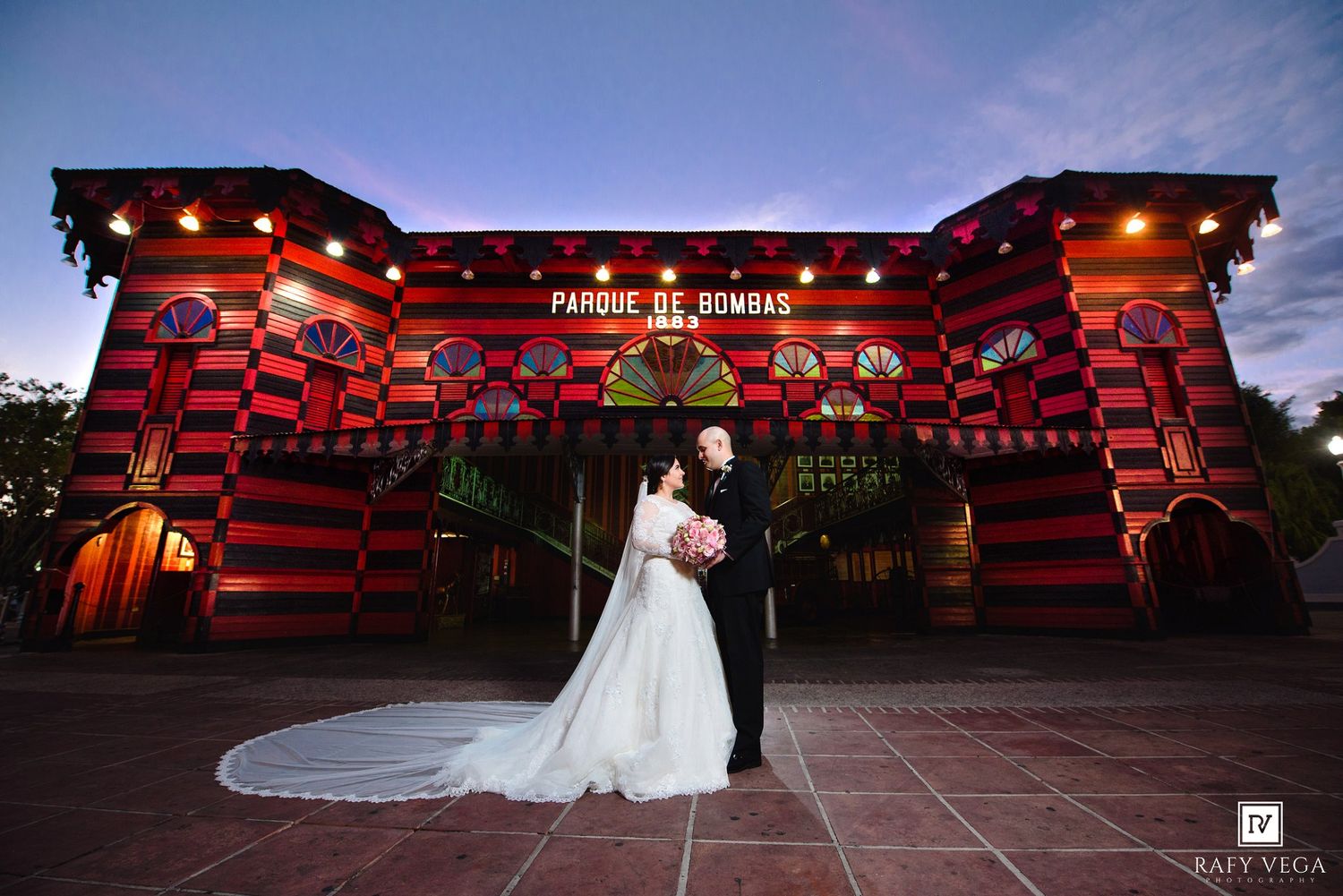Bride and groom posing in front of the iconic red Parque de Bombas building in Ponce, Puerto Rico at dusk.
