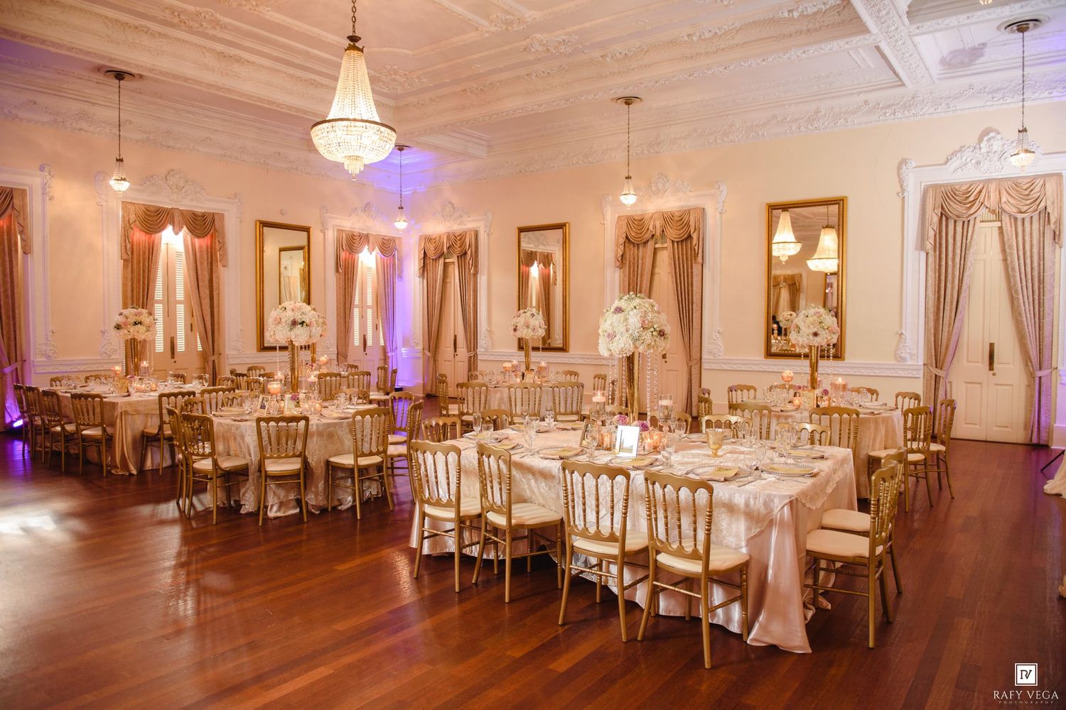 Elegant wedding reception hall with gold Chiavari chairs, white floral centerpieces, chandeliers, and warm pink lighting on hardwood floors.