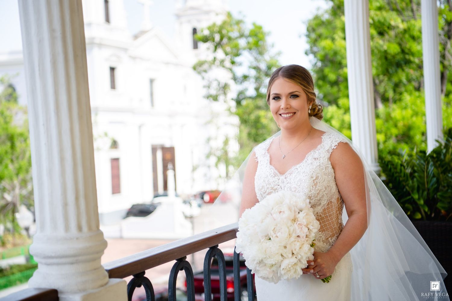 Smiling bride in lace wedding gown holds white bouquet on elegant venue balcony with columns and greenery.