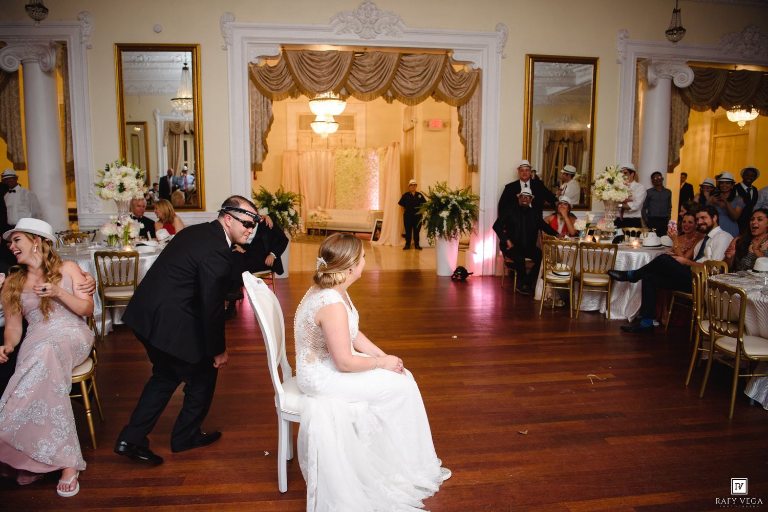 Groom dancing playfully near bride seated in chair during elegant wedding reception in ornate ballroom.
