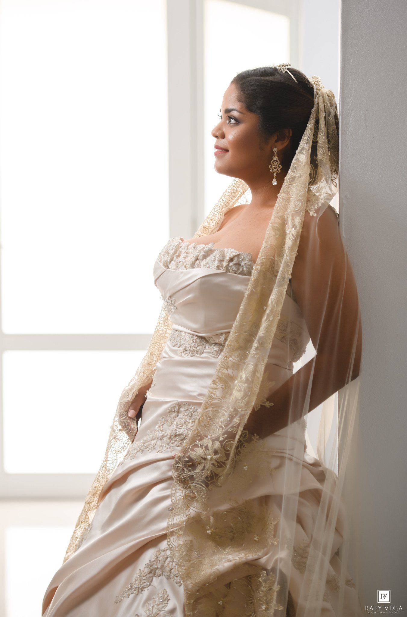 Bride in champagne satin gown with gold lace veil gazes out window in elegant bridal portrait.