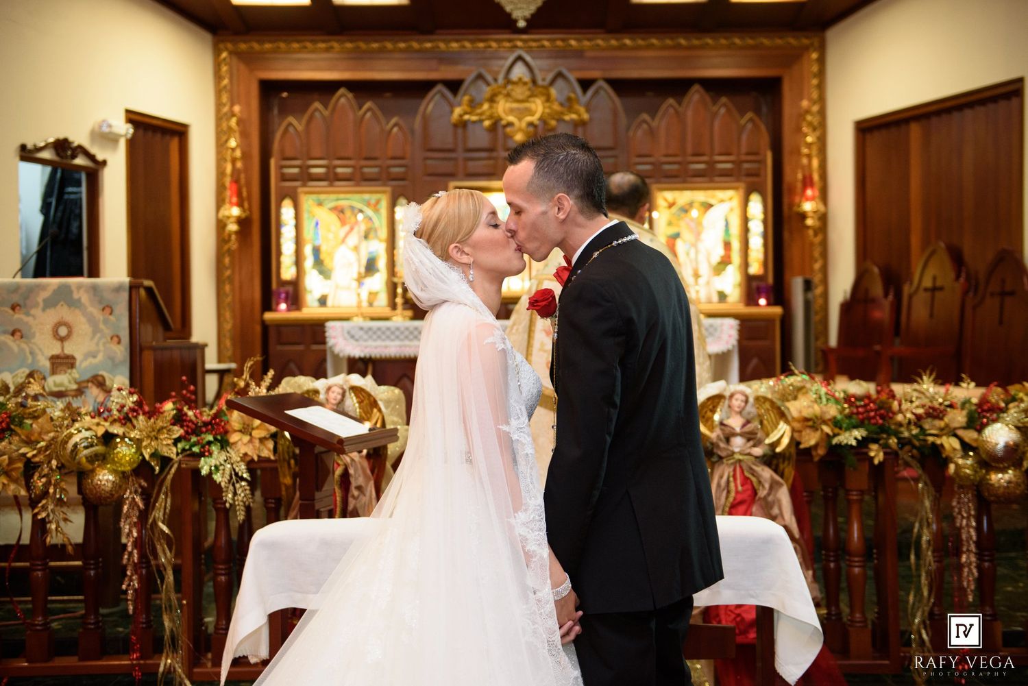 Bride and groom share first kiss at ornate church altar adorned with red and gold floral arrangements during wedding ceremony.