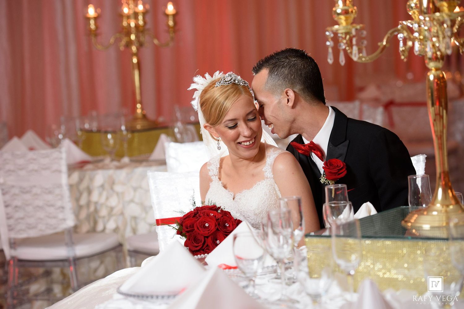 Bride and groom share a romantic moment at wedding reception table with red roses and elegant candlelit decor.