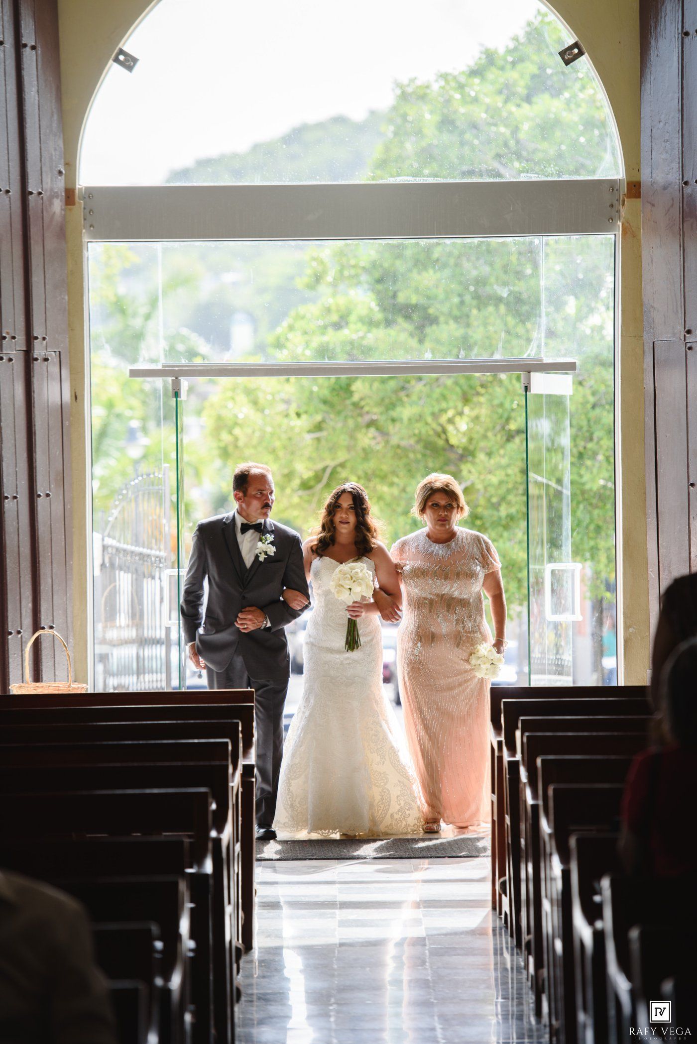 Bride walking down church aisle with parents on her wedding day, wearing a white lace gown and holding a bouquet.