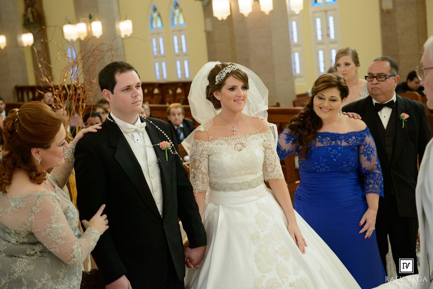 Bride in white gown and groom in tuxedo smile at wedding ceremony in church with guests in blue dress nearby.