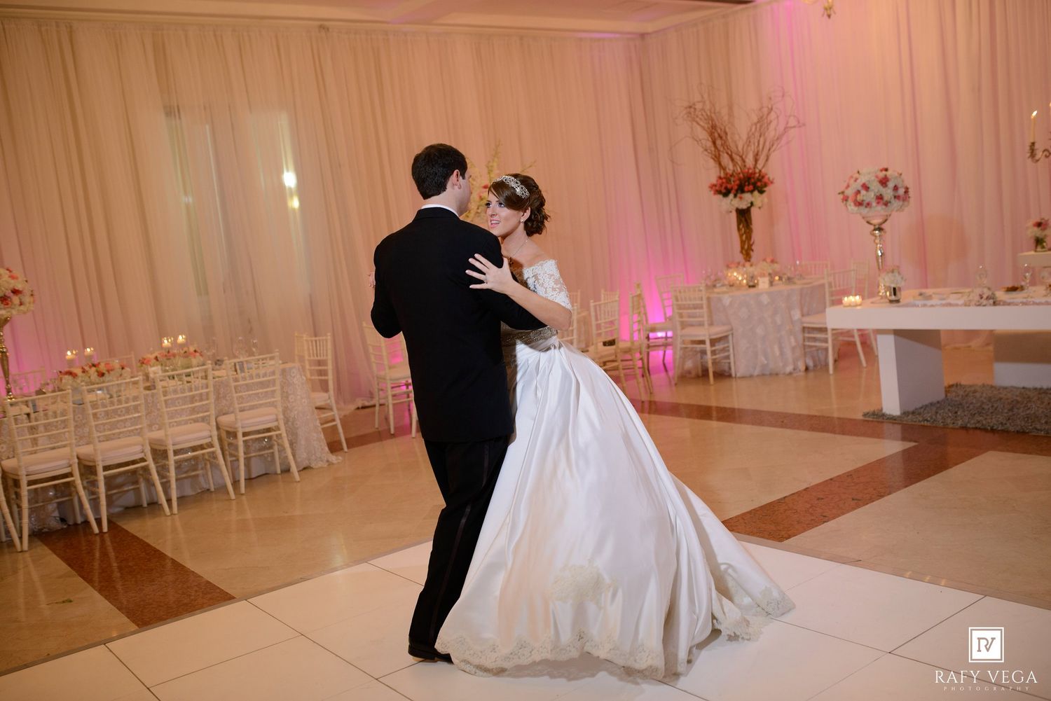 Bride and groom share their first dance on a white dance floor at an elegant wedding reception with pink lighting.