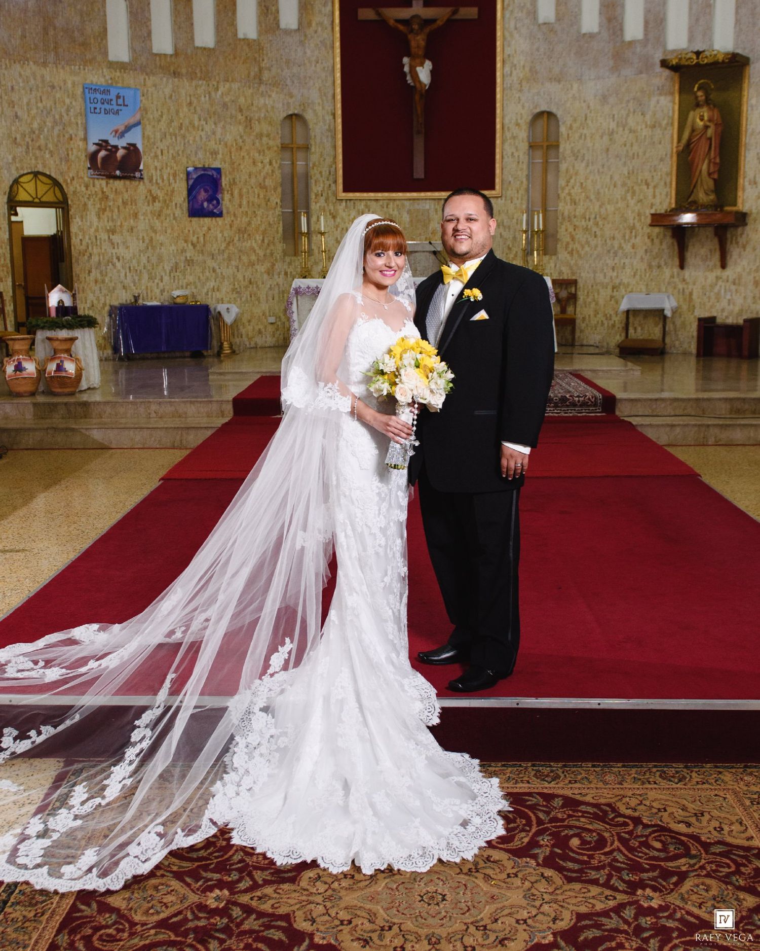 Bride and groom posing together at church altar, bride wearing lace mermaid gown with long veil and yellow bouquet.