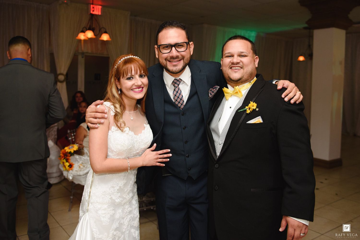 Bride and groom pose with a guest in a yellow bow tie at a wedding reception venue.