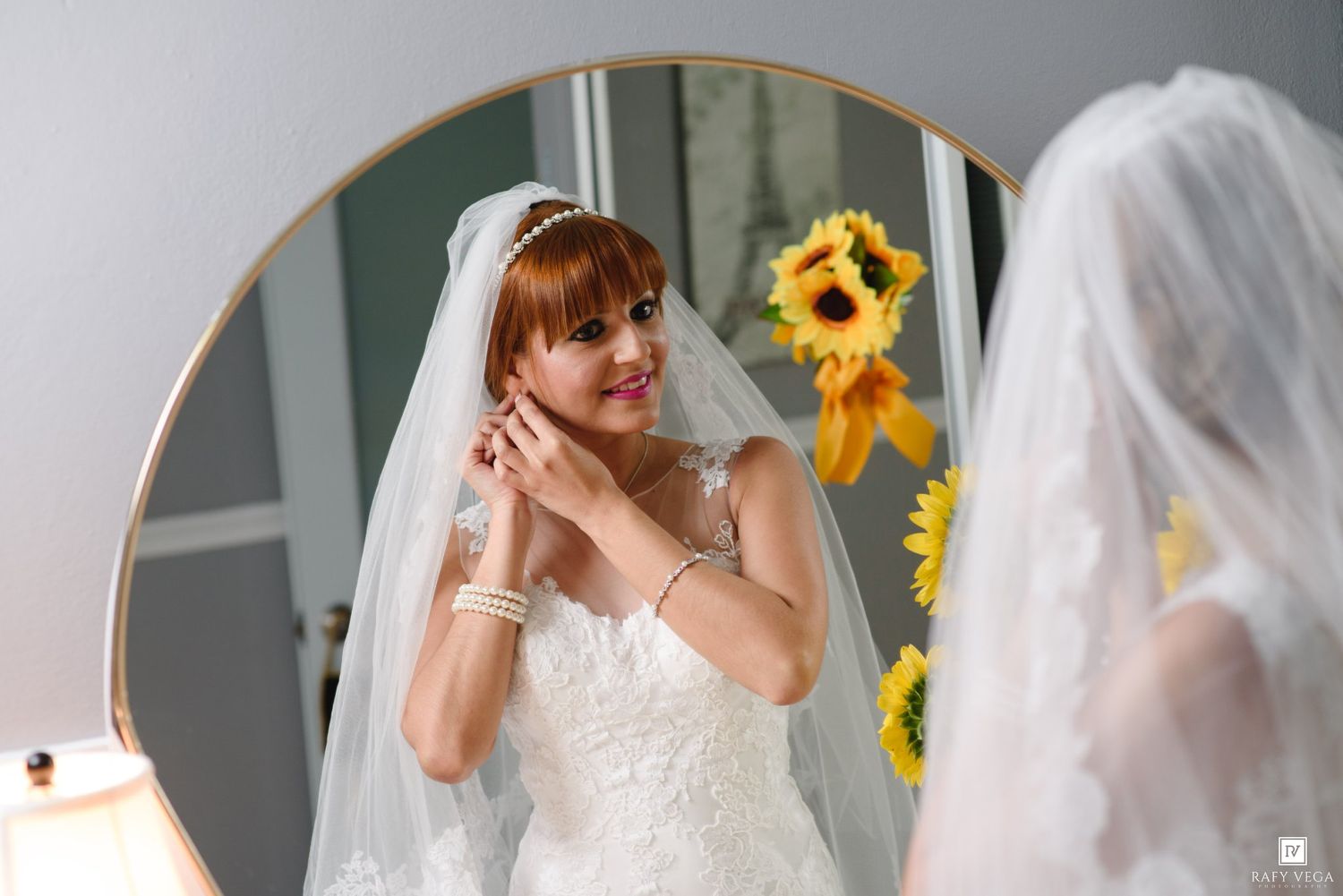 Redhead bride in white gown adjusting earrings while looking in round mirror with sunflowers.
