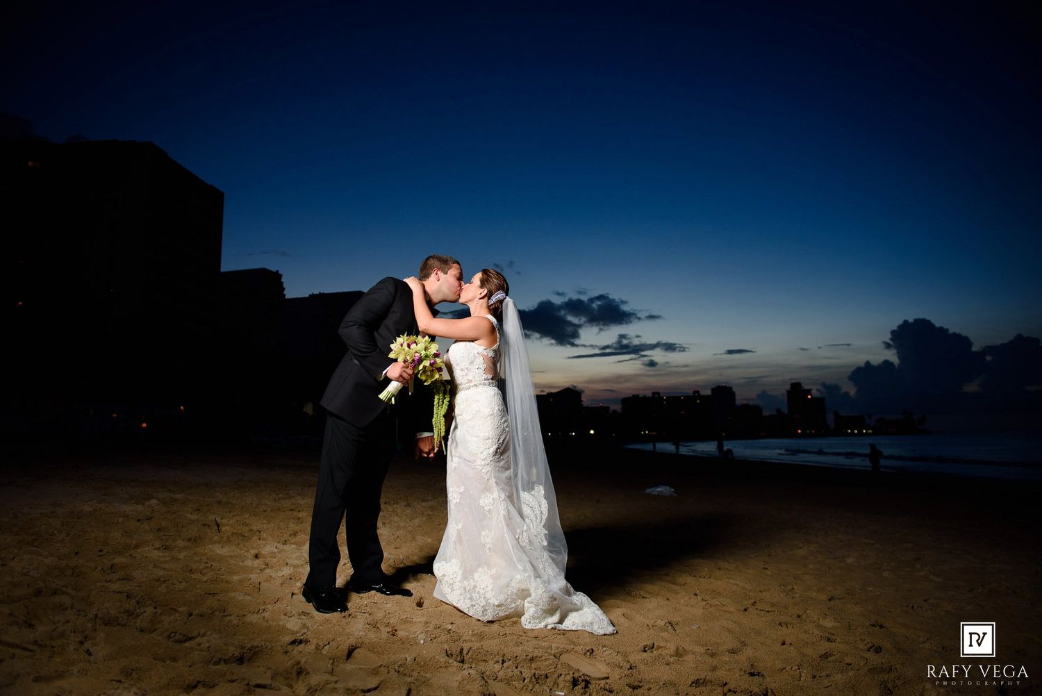 Bride and groom share a romantic kiss on a sandy beach at sunset, with a glowing twilight sky in the background.