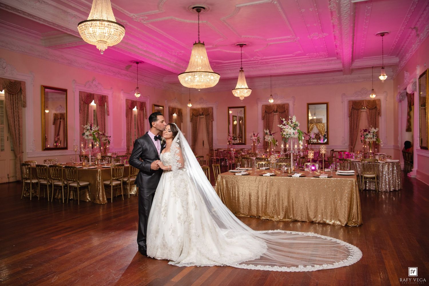 Wedding couple embraces on dance floor of luxurious pink-lit reception hall with gold tablecloths.