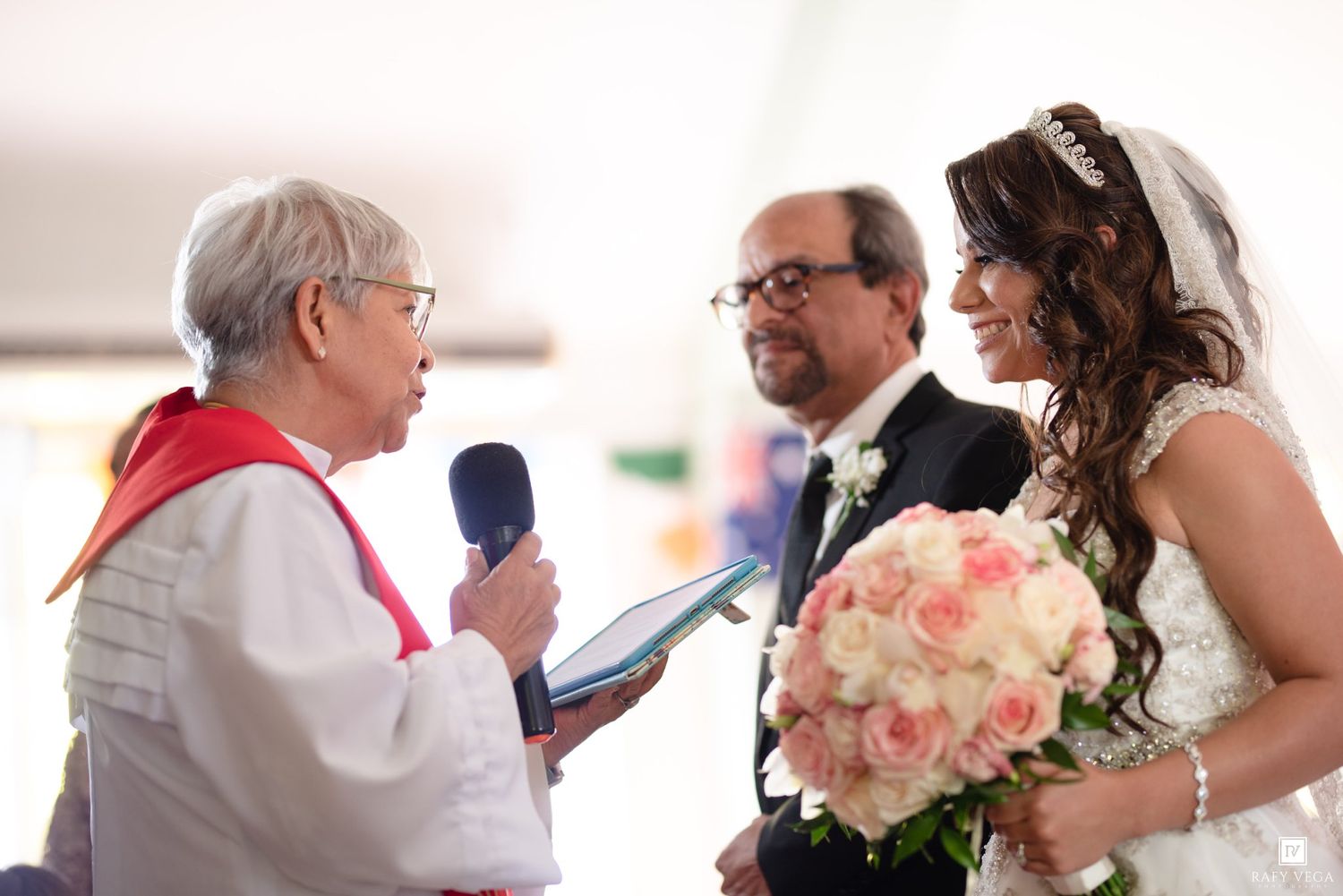 Bride holding pink roses bouquet during wedding ceremony with officiant reading vows and groom standing nearby.