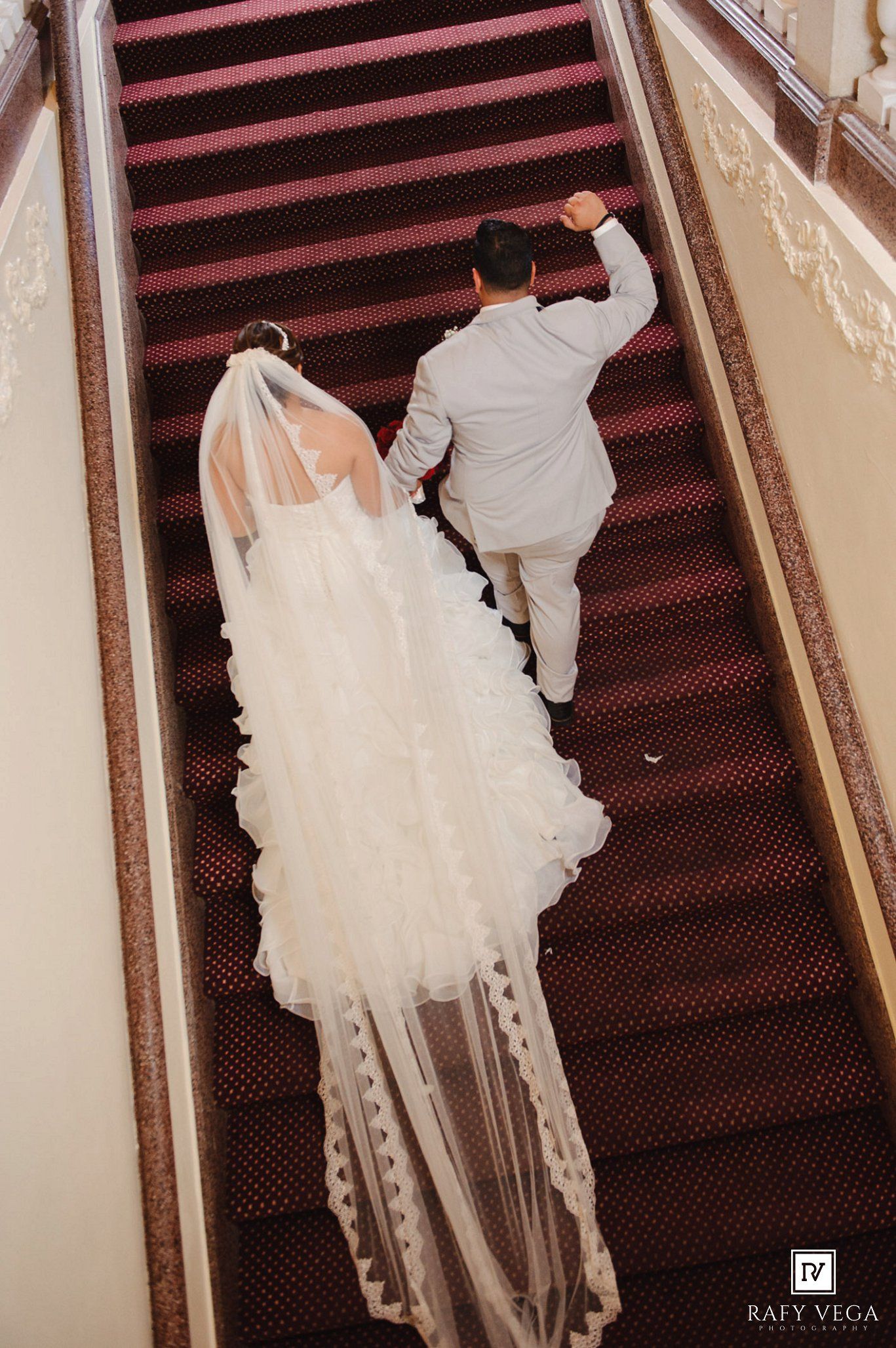 Aerial view of bride in white gown with veil and groom in gray suit walking up a red carpeted staircase.