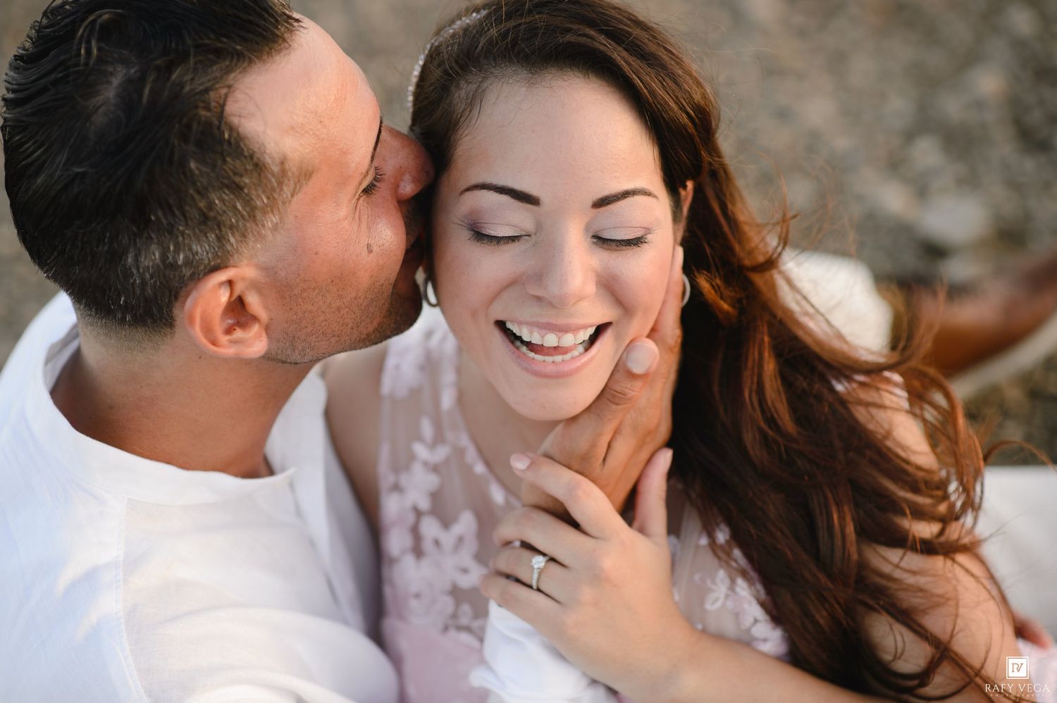 Happy couple sharing a romantic moment, woman laughing joyfully as man kisses her cheek, wearing engagement ring.