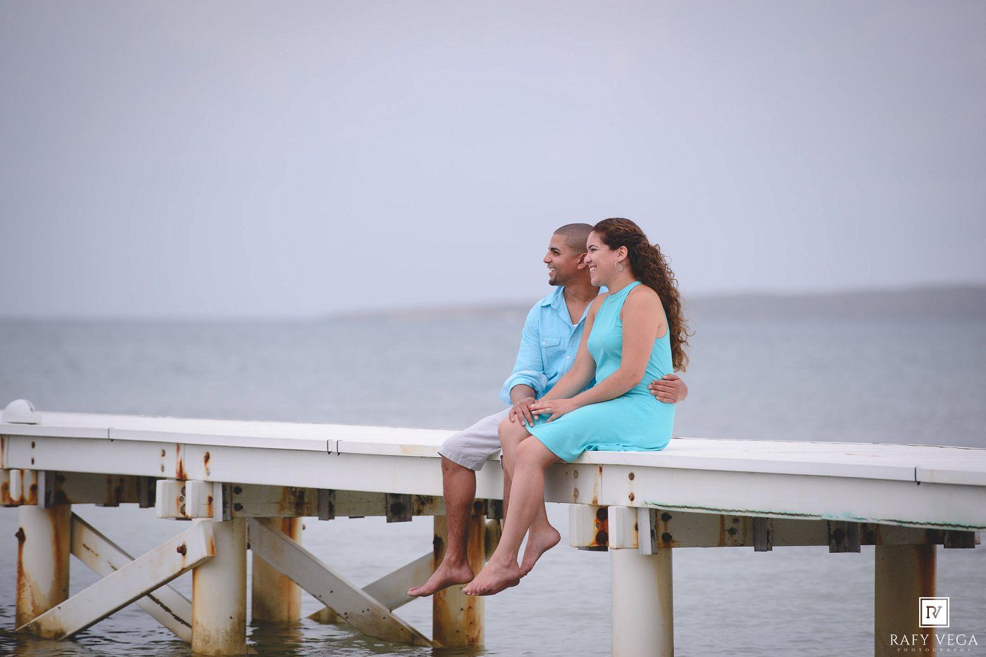 A couple sits closely together on a white pier overlooking a calm ocean on an overcast day.