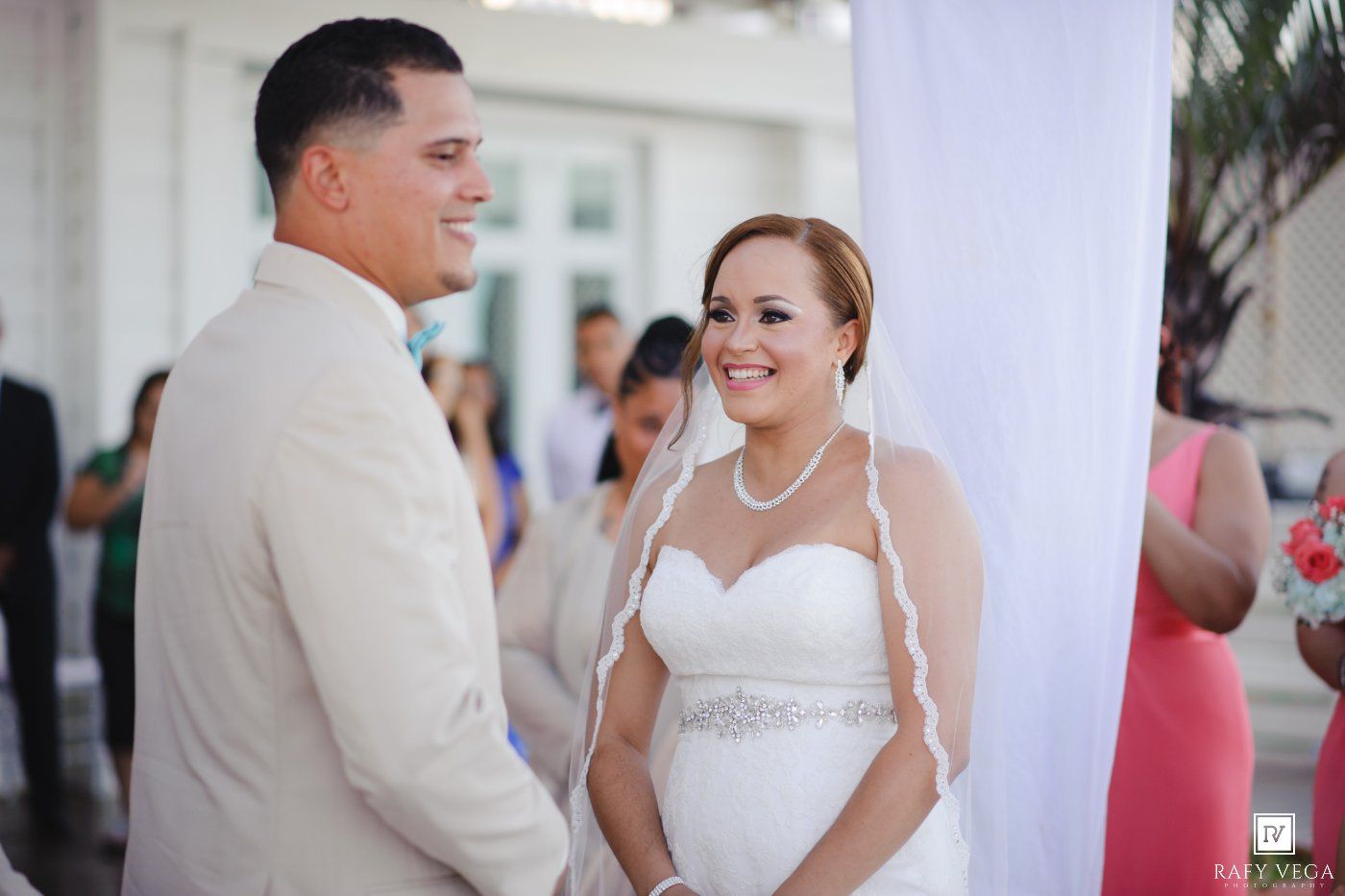 Bride and groom smiling at each other during outdoor wedding ceremony, bride wearing strapless gown with veil.