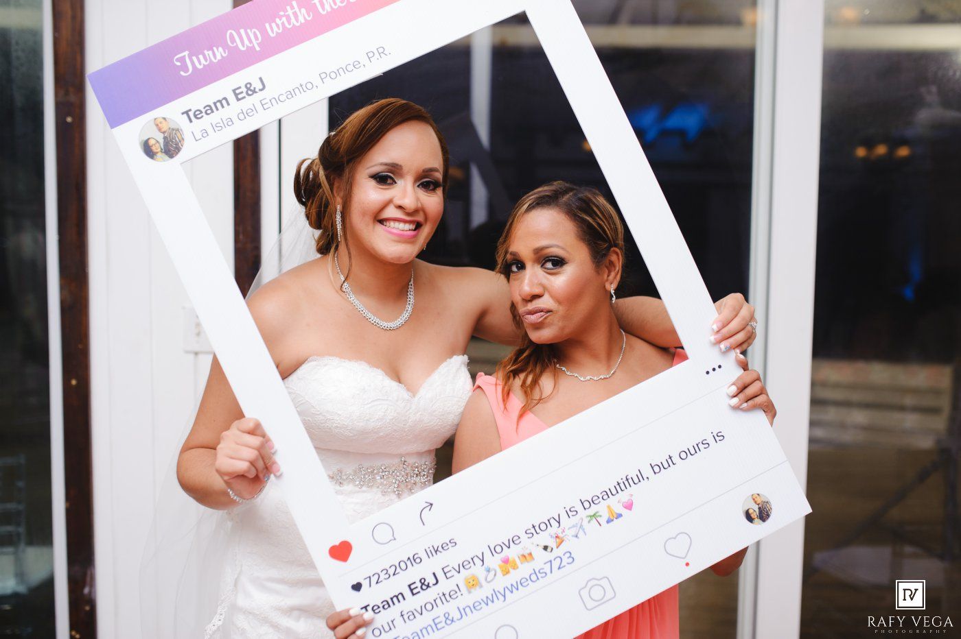 Bride and bridesmaid smiling together holding an oversized Instagram photo frame prop at a wedding reception.