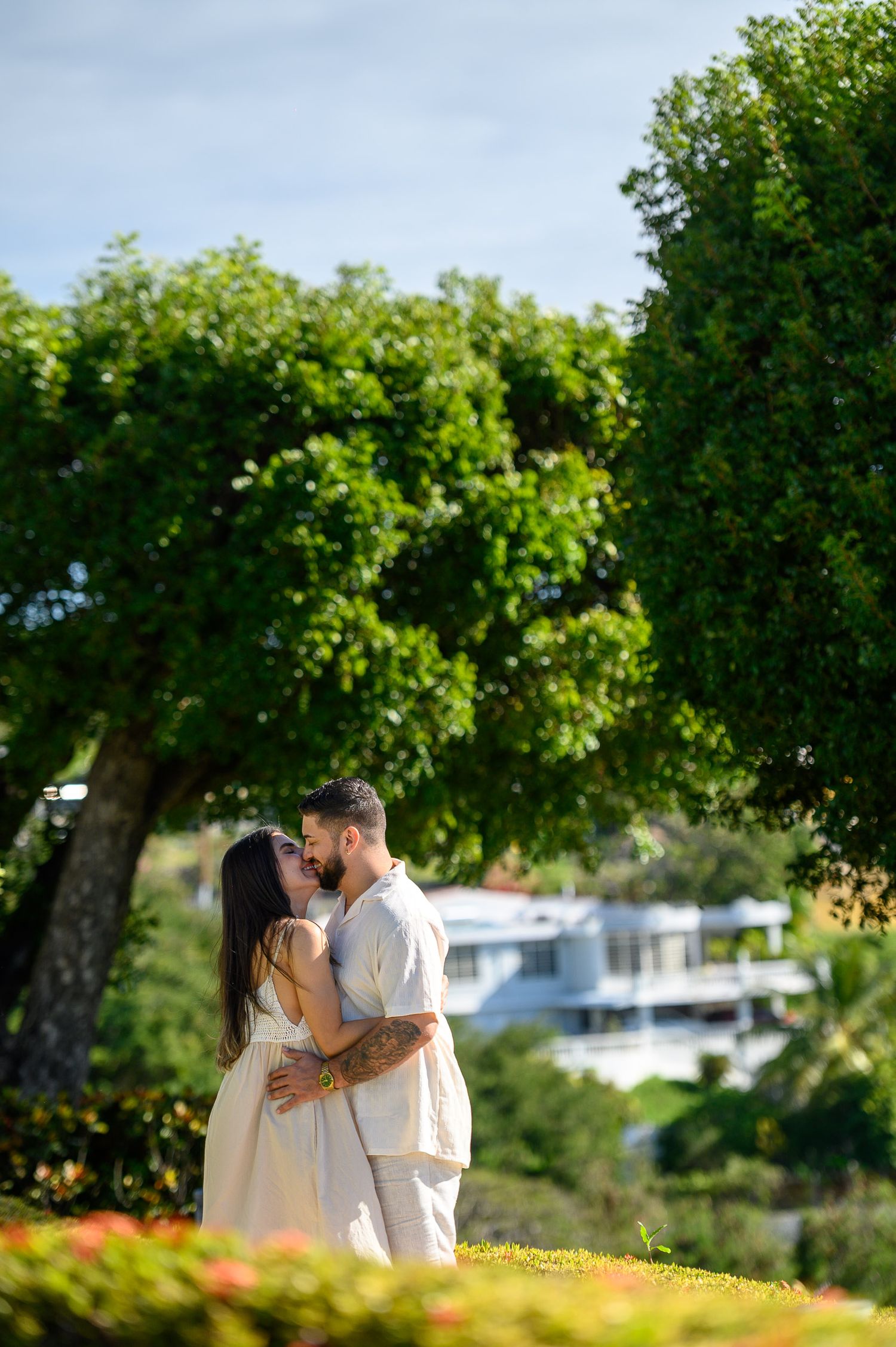 Couple sharing a romantic kiss in a lush green garden with trees and a white building in the background.