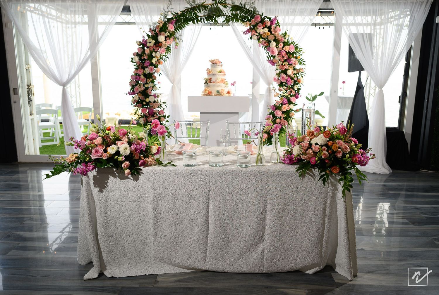 Elegant wedding reception table with pink and peach floral arrangements, a floral arch, and a tiered cake beneath sheer white drapes.