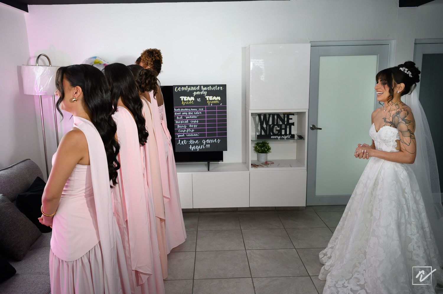 Bridesmaids in blush pink dresses turn to look at a bride in a white lace gown with an updo in a bright room with a wine night sign.