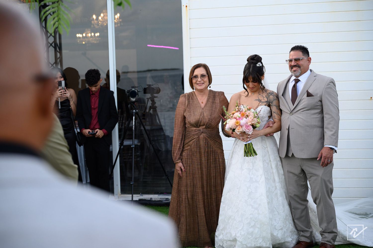 Bride in a white gown holding a pink bouquet stands arm in arm with a woman in a brown dress and a man in a gray suit at an outdoor wedding.