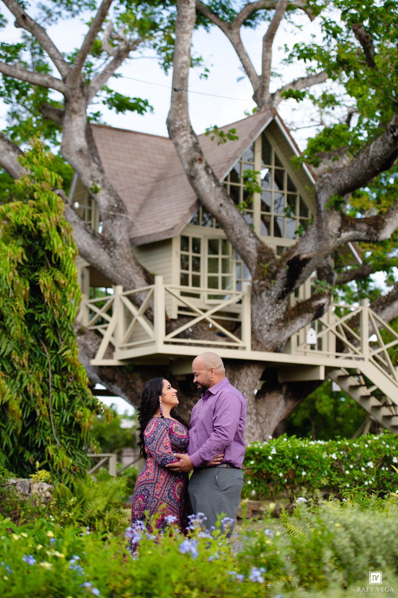 Romantic couple embracing in front of a charming treehouse surrounded by blooming flowers and large trees.