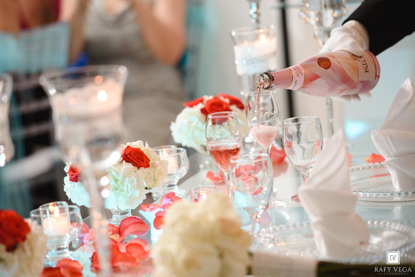 Waiter pouring rose champagne into crystal glasses at elegant wedding reception with floral centerpieces and candles.