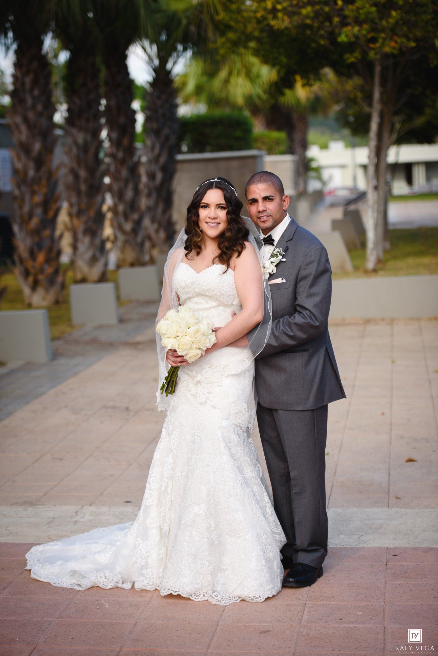 Bride in white lace gown holding bouquet stands with groom in gray suit outdoors near palm trees on their wedding day.