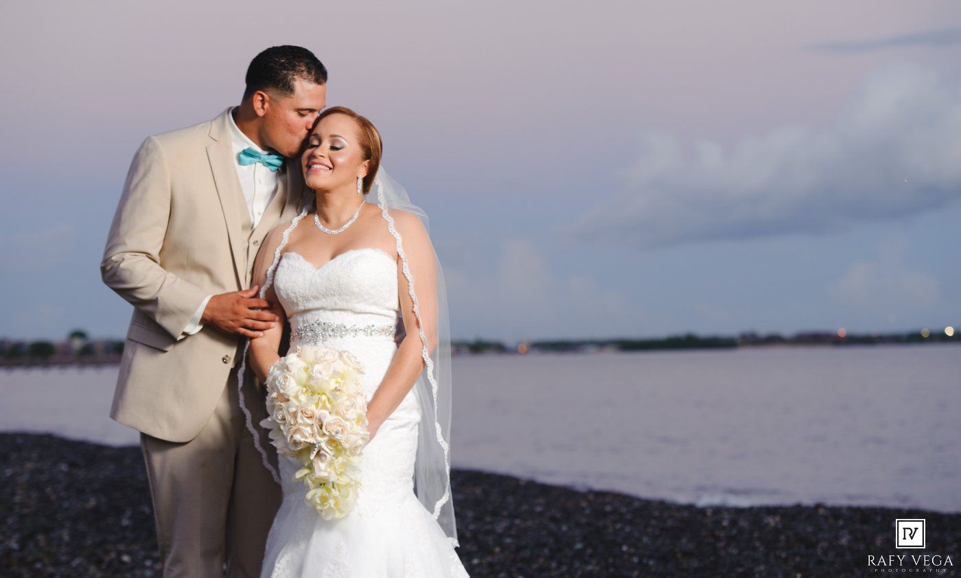 Couple embraces on a beach at sunset, groom in tan suit with teal bow tie, bride in white gown with bouquet.