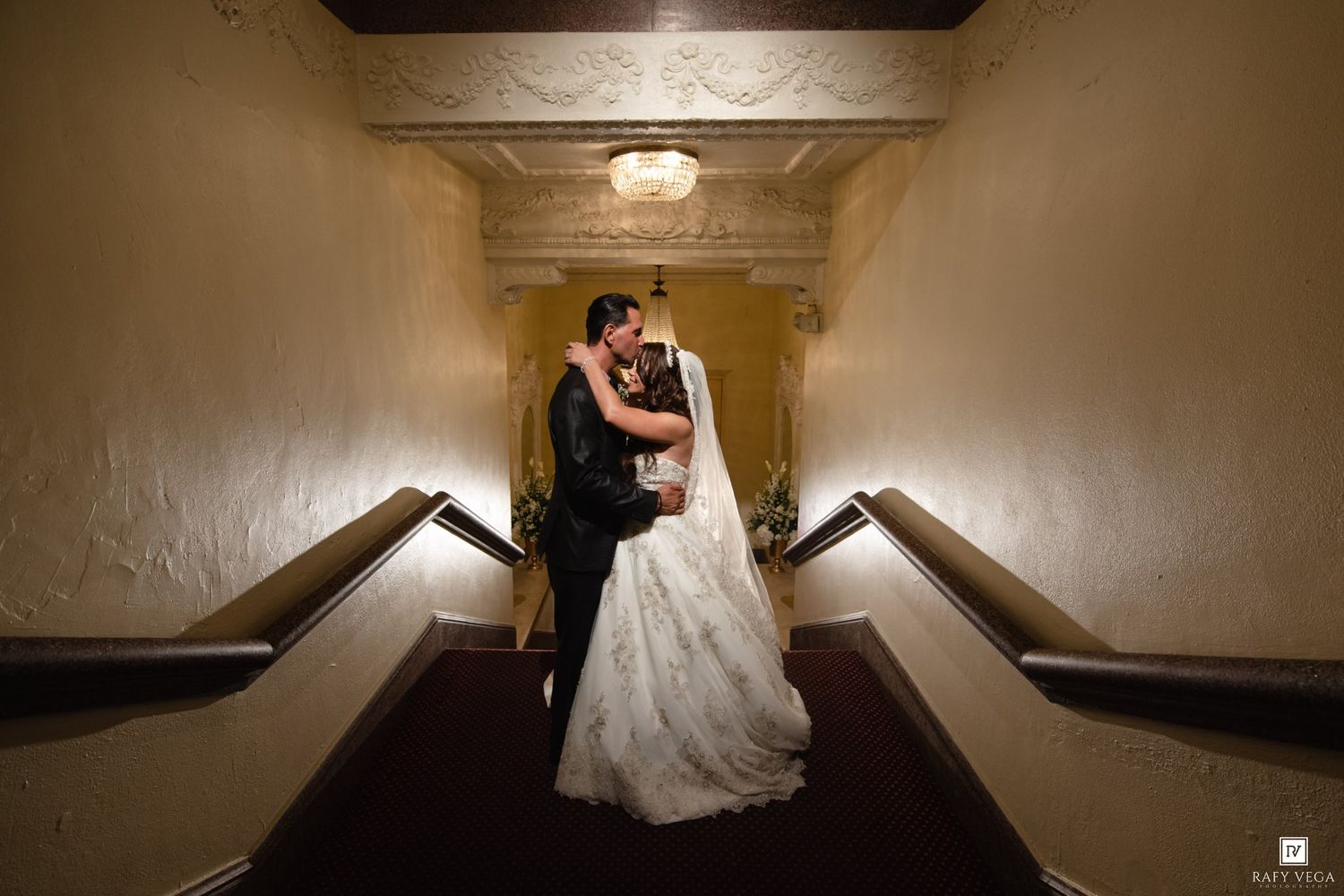 Bride and groom share a romantic kiss on elegant staircase in ornate wedding venue hallway.